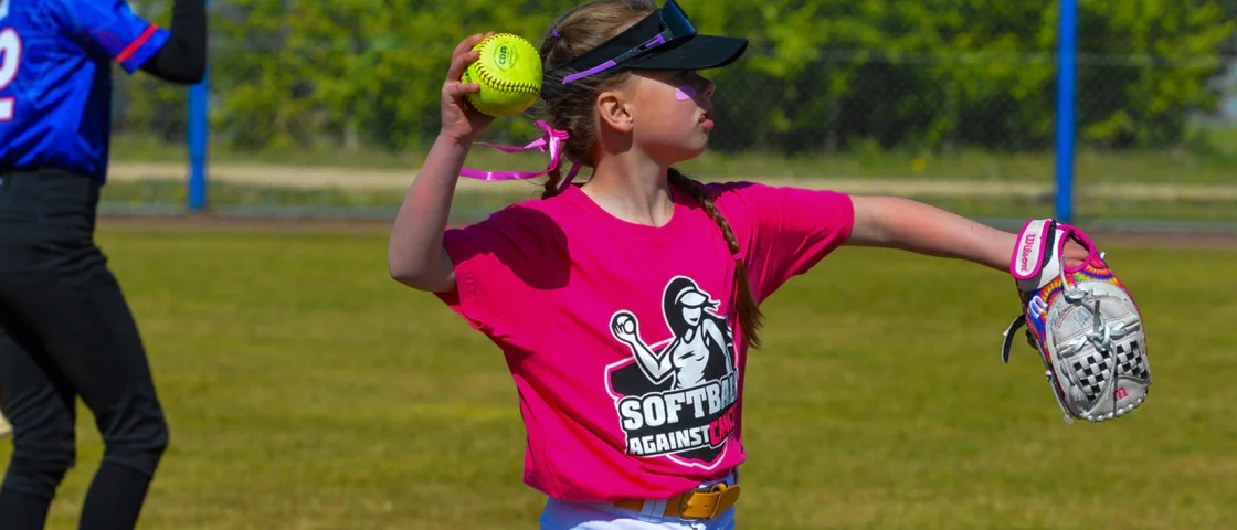 Jonge slagvrouwen in roze, blauwe en groene shirts staan met hun knuppels in de rij langs het hek tijdens Softball Against Cancer