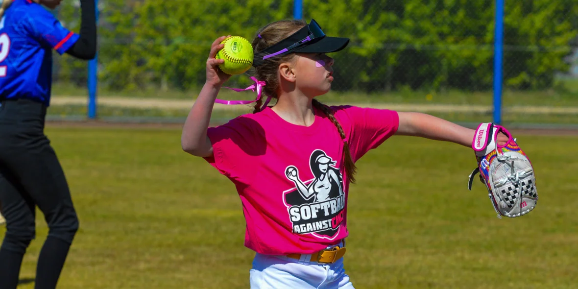 Jonge slagvrouwen in roze, blauwe en groene shirts staan met hun knuppels in de rij langs het hek tijdens Softball Against Cancer