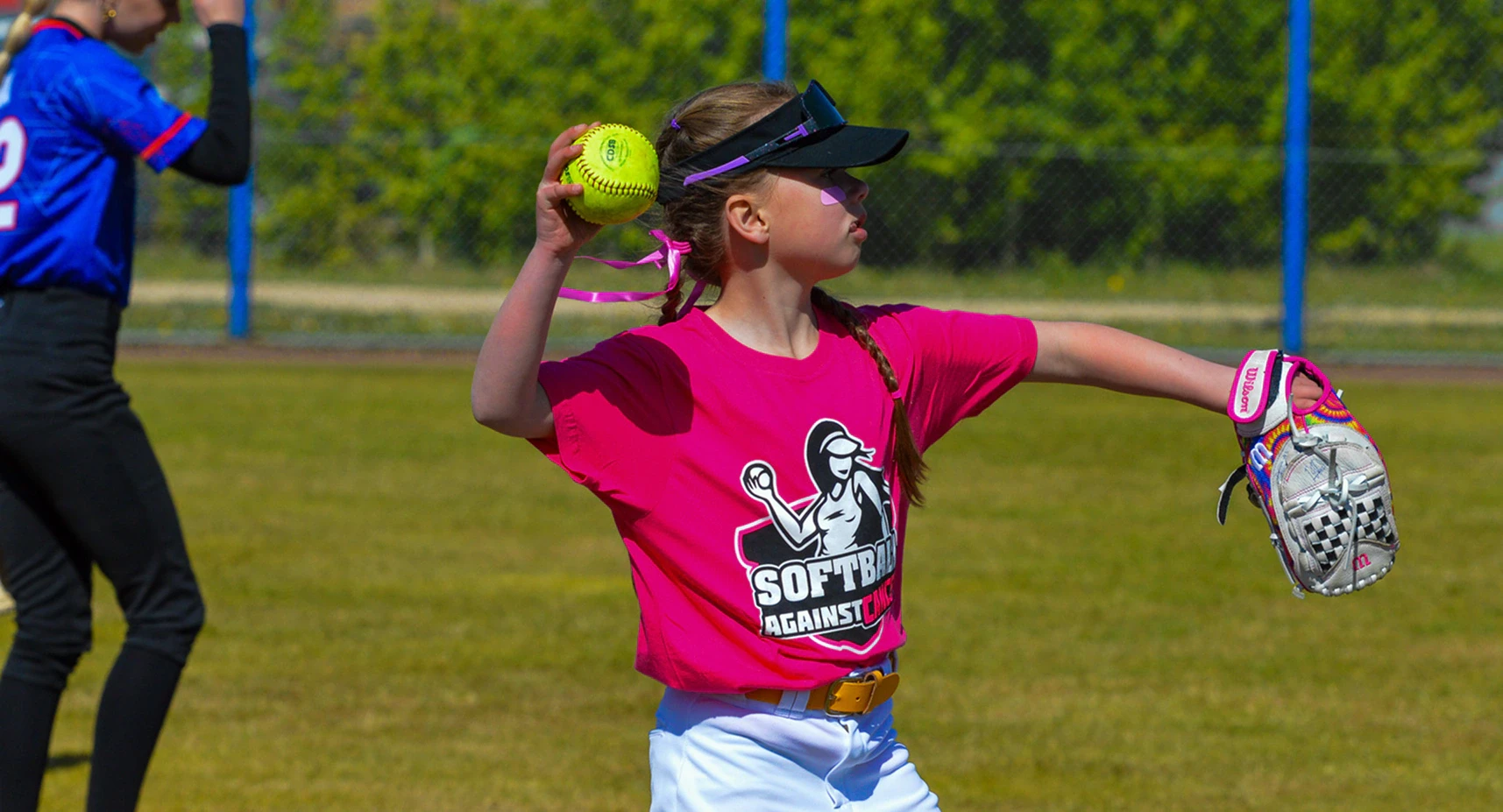 Jonge slagvrouwen in roze, blauwe en groene shirts staan met hun knuppels in de rij langs het hek tijdens Softball Against Cancer
