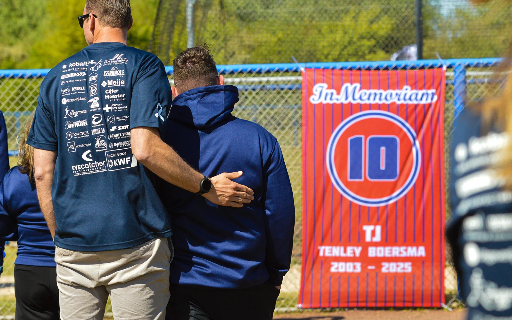 Twee mannen staan voor de In Memoriam banner van Tenley Boersma rugnummer 10, 2003-2025, tijdens Softball Against Cancer