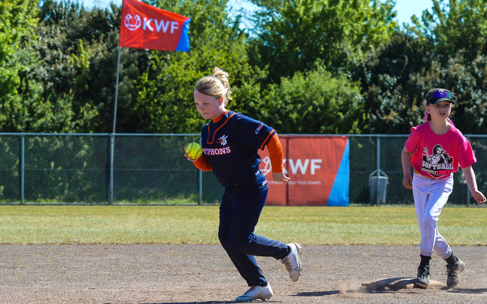 Jonge softbalspeelster van de Gryphons rent met de bal langs het honk, met op de achtergrond de KWF-vlag van het benefiettoernooi