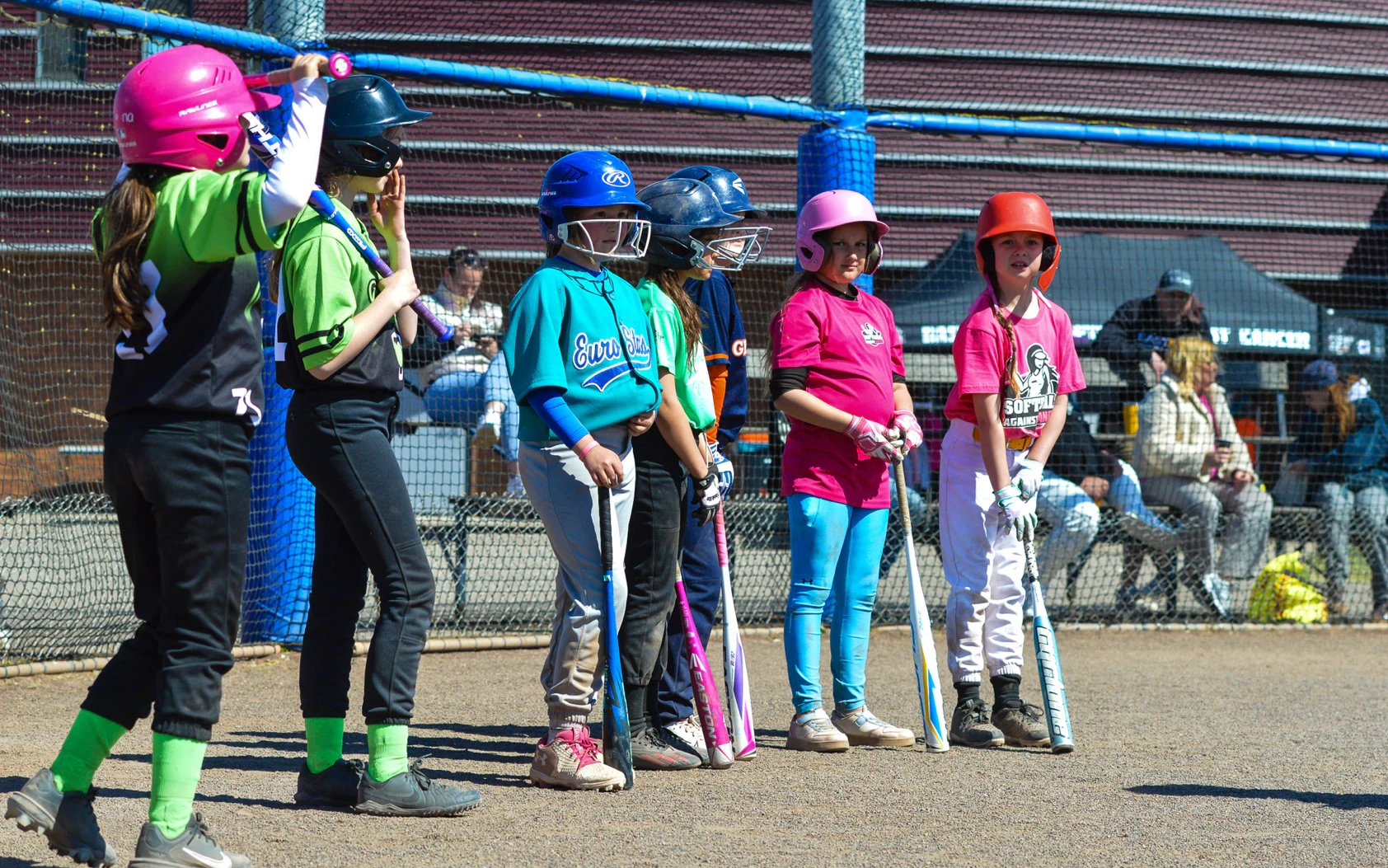 Jonge slagvrouwen in roze, blauwe en groene shirts staan met hun knuppels in de rij langs het hek tijdens Softball Against Cancer