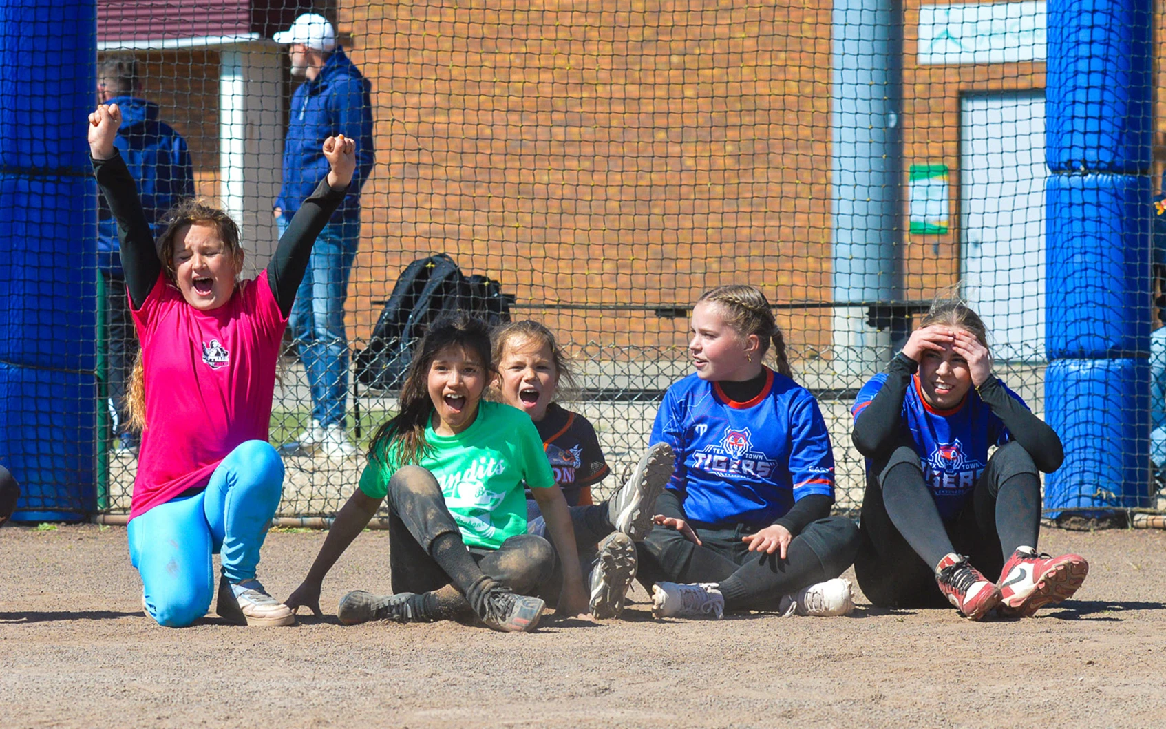 Vijf jonge softbalspeelsters in verschillende teamshirts juichen en reageren langs de zijlijn van het softbalveld