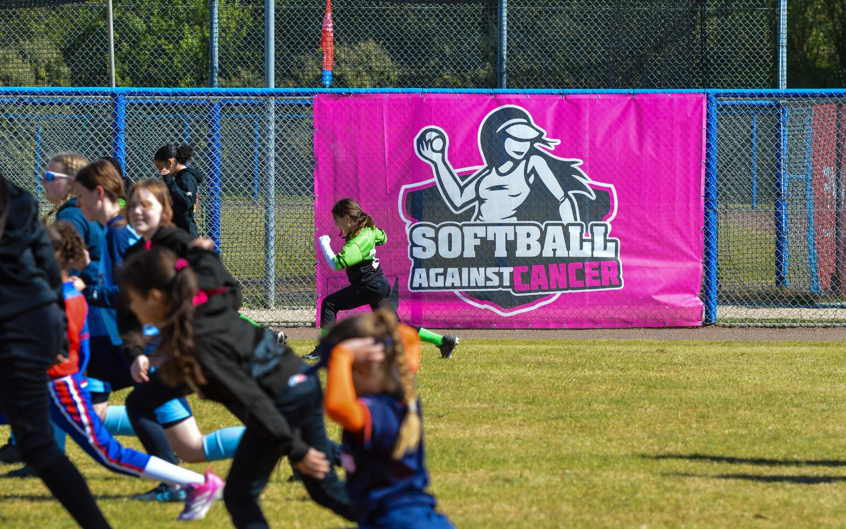 Jonge speelsters rennen langs de roze Softball Against Cancer banner tijdens de warming-up van het benefiettoernooi