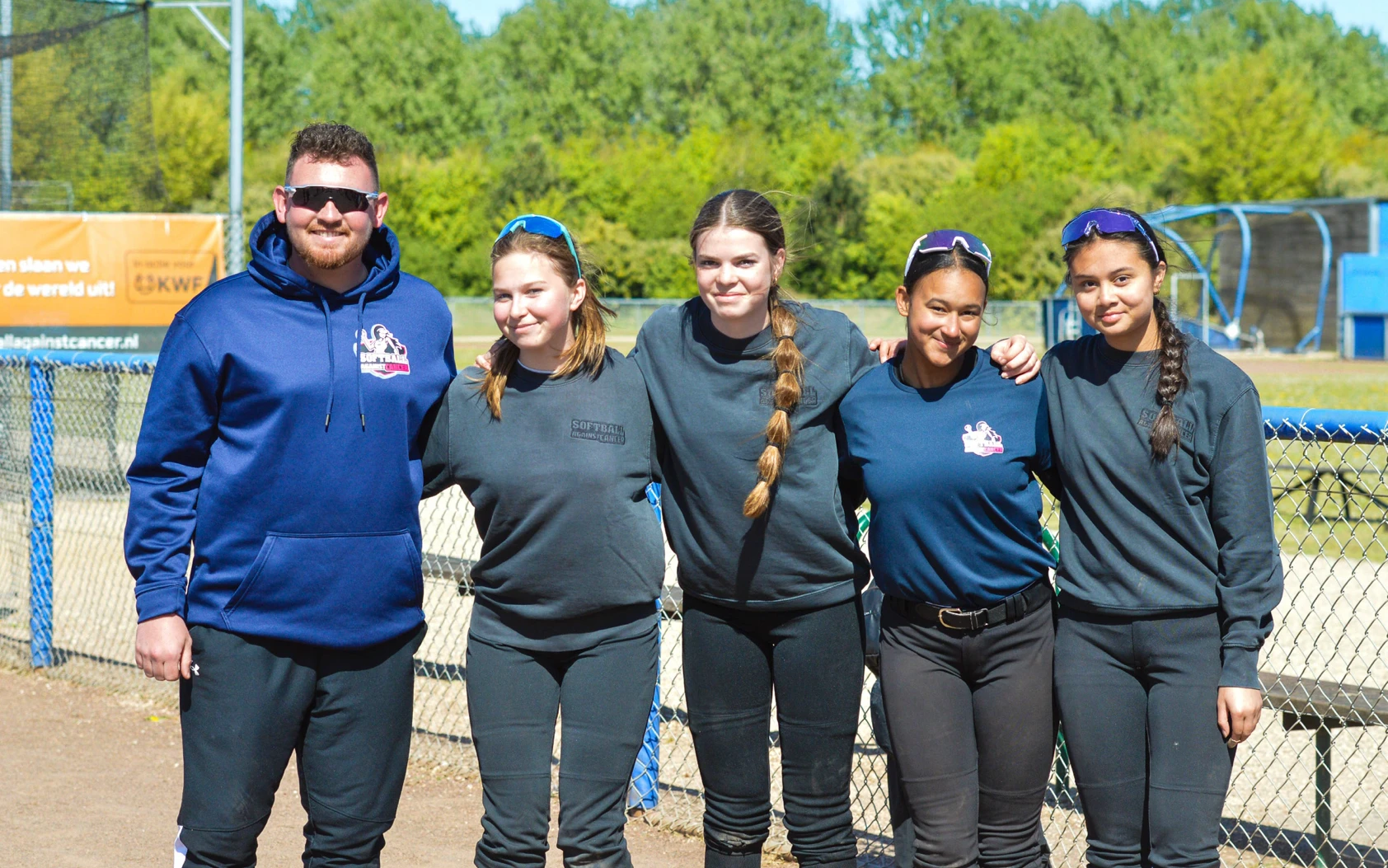 Coach poseert met vier jonge softbalspeelsters in donkere outfits voor het hek van het softbalveld tijdens Softball Against Cancer
