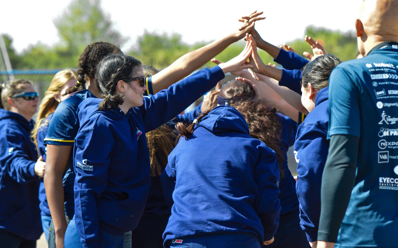 Softbalteam in blauwe hoodies steekt de handen omhoog in een huddle voor aanvang van de wedstrijd Softball Against Cancer