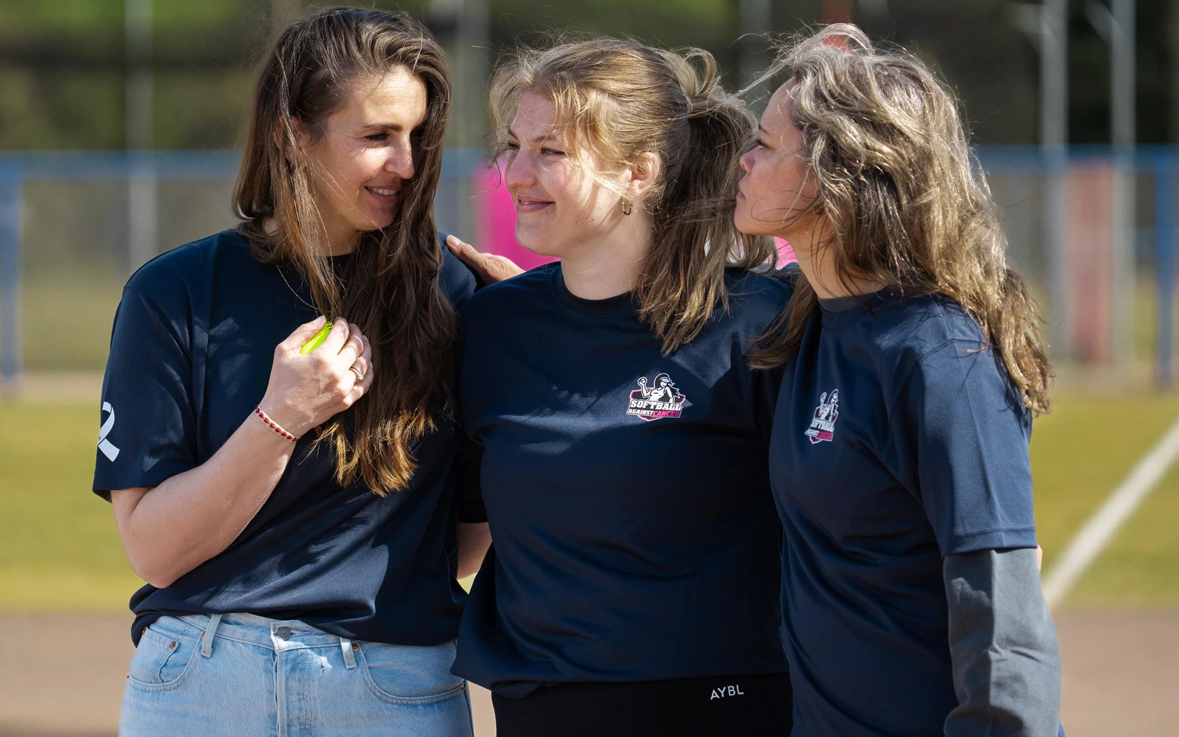 Drie vrouwen van de organisatie in Softball Against Cancer shirts staan glimlachend naast elkaar op het softbalveld