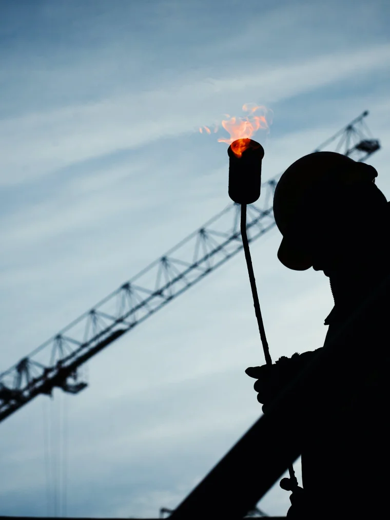 Afbeelding van een man met een helm en een vlammend iets vast op een bouwplaats. Er staat een kraan op de achtergrond
