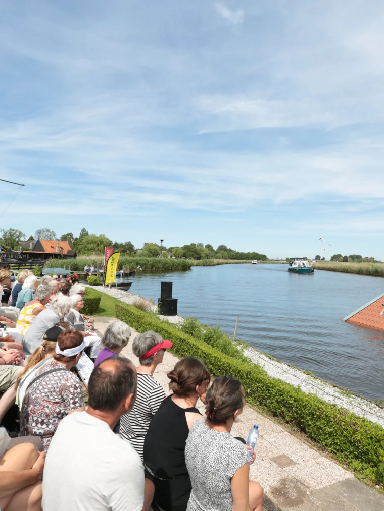 Afbeelding van publiek aan de rand van water waarin een huis staat. Alleen het dak steekt uit en er liggen 4 mensen op