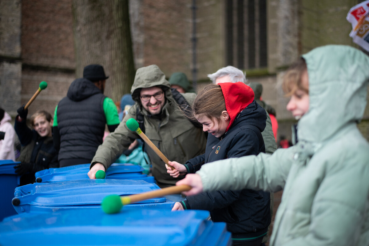 Groep mensen slaan met 'drumsticks' (stokken met groene bollen aan de uiteindes) op vuilnisbakken