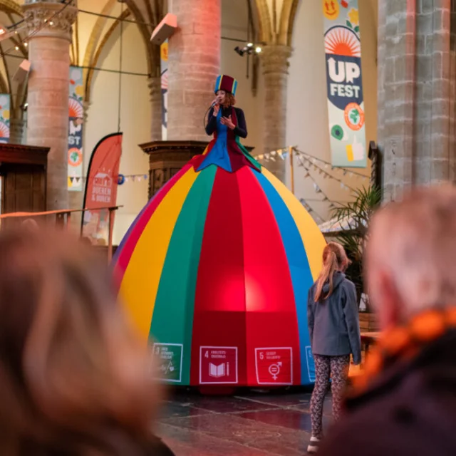 Vrouw op stelten met een enorme rok in de kleuren van een circustent is aan het zingen