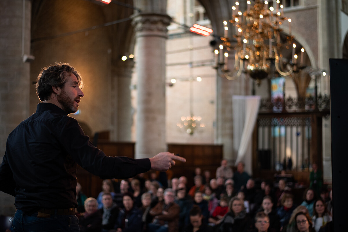 Man, zwart gekleed, doet een toespraak voor een groep mensen. Het lijkt in een kerk te zijn
